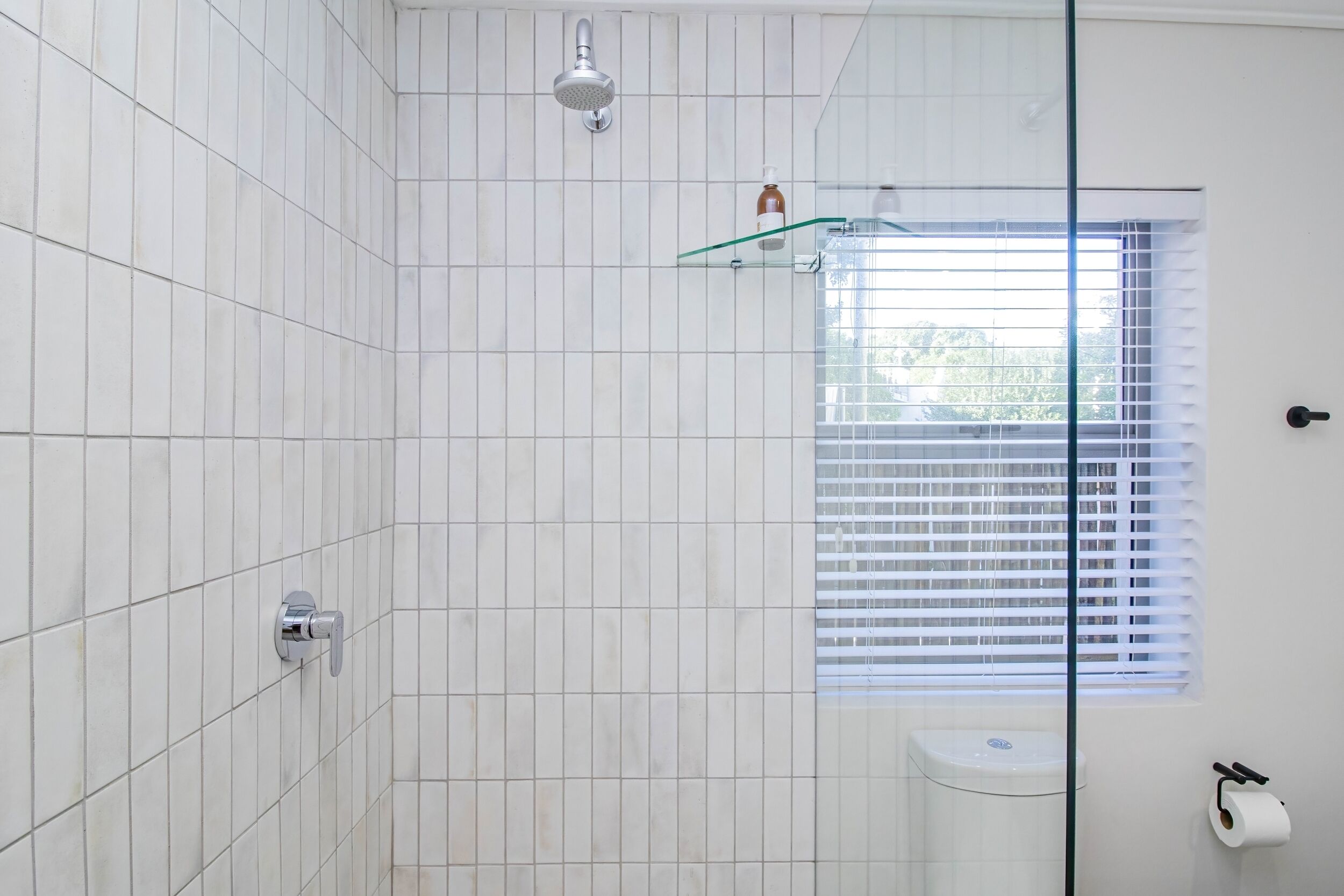 A bathroom with white tiled walls, a glass shower enclosure, a toilet, and a window with blinds.