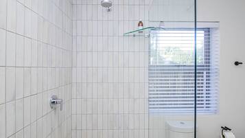 A bathroom with white tiled walls, a glass shower enclosure, a toilet, and a window with blinds.