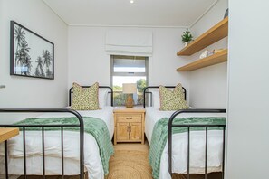 A bedroom with two beds, a window, a bedside table, a lamp, a shelf with decorative items, and a framed picture on the wall. - Pikkewyntjie | Sea view | Small families (Vermont)