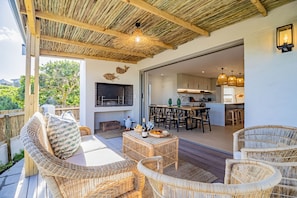 An outdoor patio with wicker furniture, a grill, and a dining area with a kitchen in the background.