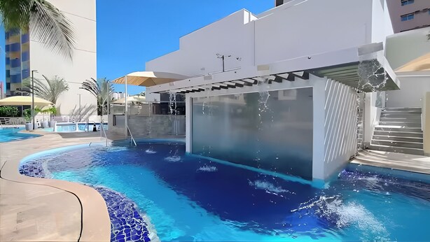 Zona de piscina moderna de hotel con una piscina azul cristalina, un jacuzzi con paredes de vidrio y una escalera que lleva a un nivel superior.
