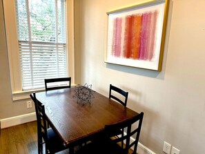 Un área de comedor con una mesa de madera y cuatro sillas, una ventana con persianas, y un cuadro abstracto enmarcado en la pared.