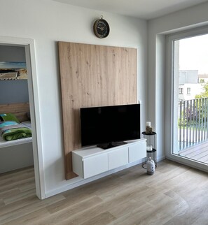Un salon moderne avec un téléviseur à écran plat posé sur un meuble blanc, un mur d’accent en bois et une grande fenêtre donnant sur un balcon.