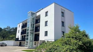 A modern multi-story building with a glass elevator shaft, a car parked in front, and lush greenery around. - Seaside 53° FeWo 7 - Apartment close to the beach with sea view and balcony in (Cuxhaven)