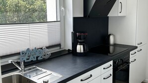 A modern kitchen with a built-in stove, oven, and coffee maker, white cabinets, and a window with blinds. - Seaside 53° FeWo 7 - Apartment close to the beach with sea view and balcony in (Cuxhaven)