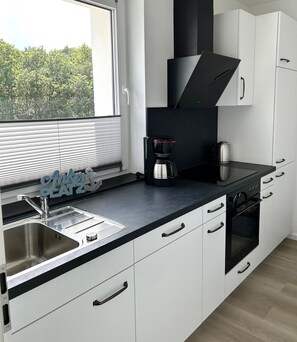 A modern kitchen with a built-in stove, oven, and coffee maker, white cabinets, and a window with blinds.