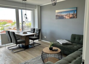 Un salon moderne comprenant un coin repas, un canapé vert, une table basse ronde agrémentée d’une plante et un tableau représentant une scène de plage accroché au mur.
