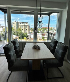 Un espace de salle à manger moderne, doté d’une grande table, de quatre chaises rembourrées et offrant une vue sur le paysage urbain grâce à de larges fenêtres.