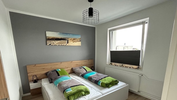 A modern bedroom with a wooden headboard, a large window, and a beach-themed painting on the wall. - Seaside 53° FeWo 7 - Apartment close to the beach with sea view and balcony in (Cuxhaven)