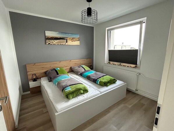 A modern bedroom with a wooden headboard, a large window, and a beach-themed painting on the wall.