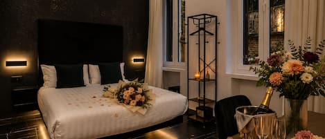 A modern hotel room with a large bed, bedside tables, a desk with a vase of flowers, a bottle of champagne, and a cake.