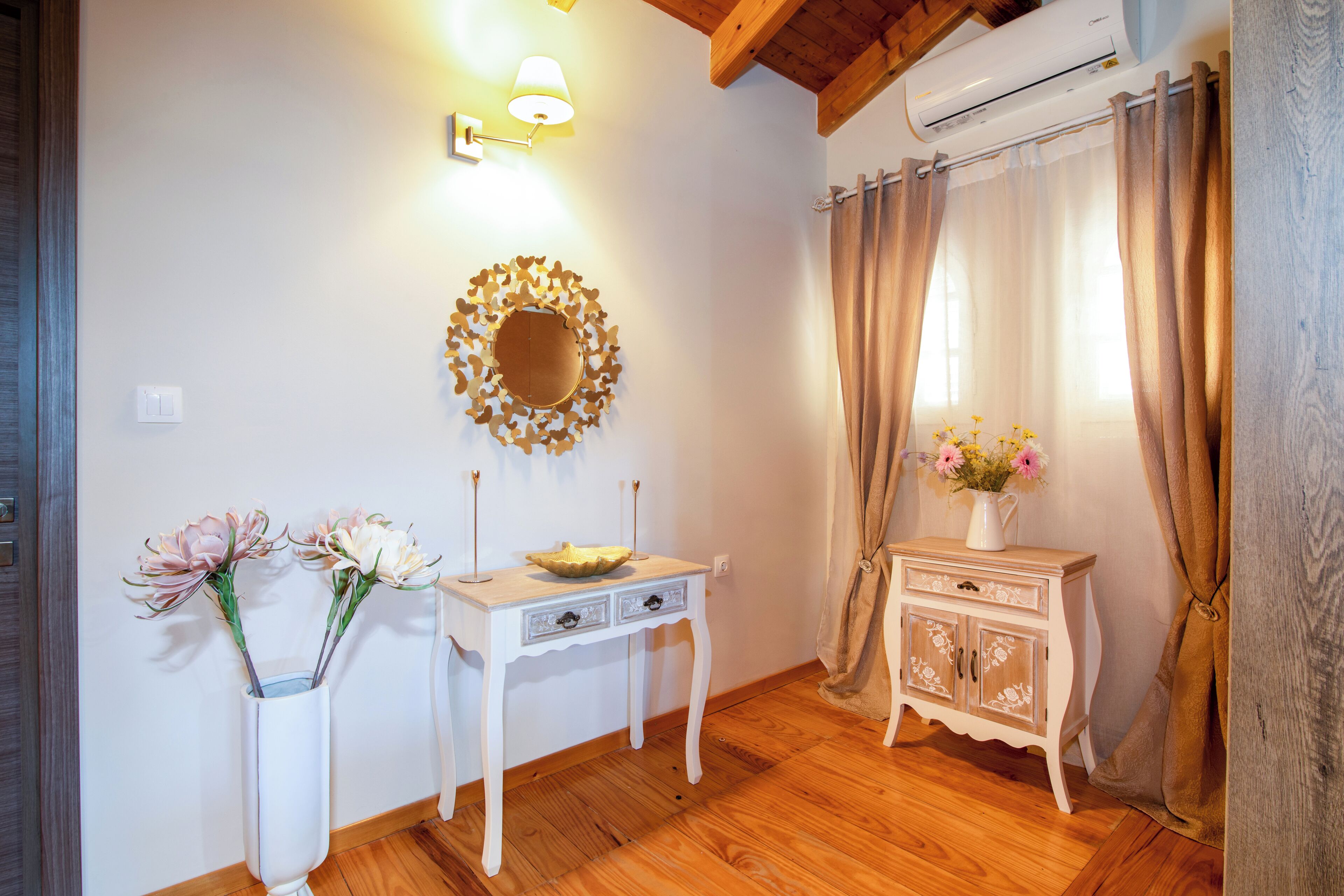 A room with a wooden floor, a white console table with drawers, a decorative mirror, a vase with flowers, and a cabinet with floral patterns.