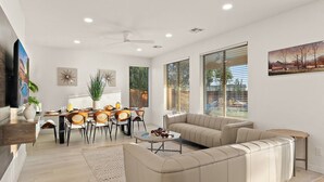 A modern living room with a beige sofa, dining table, and chairs. There is a TV mounted on the wall, a ceiling fan, and large windows with blinds.