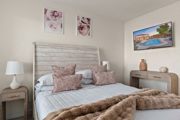 A bedroom with a wooden headboard, a bed with patterned pillows, a fluffy brown throw, a nightstand with a lamp, and a painting of a pool on the wall.