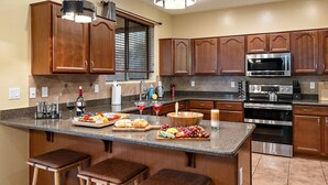 A modern kitchen with wooden cabinets, a large island with bar stools, stainless steel appliances, and a window with blinds. - Chill & Thrill | Pool, Hot Tub • Family Game Villa (Casa Grande)