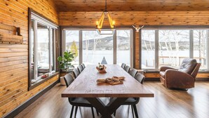 Une pièce aux murs lambrés, avec une grande table à manger, des chaises et une vue sur un lac, visible à travers de grandes fenêtres.
