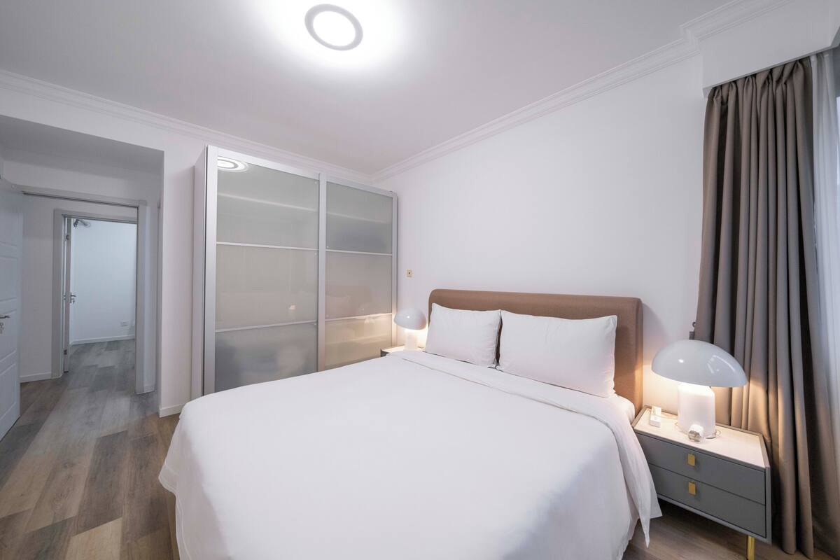 A modern bedroom with a large bed, a nightstand with a lamp, a wardrobe with sliding doors, and a door leading to another room.