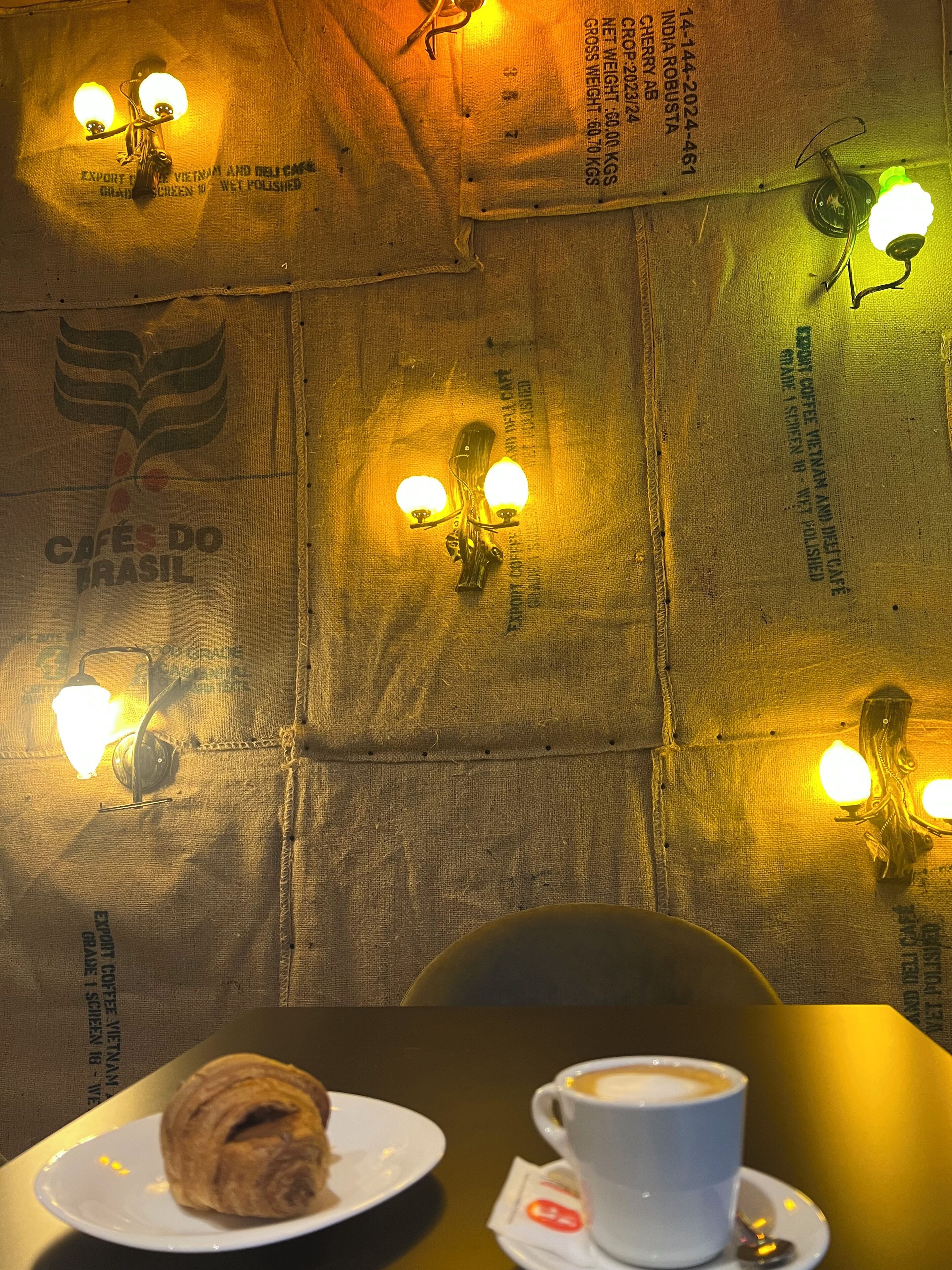 Une table avec un croissant et une tasse de café, un mur avec des luminaires et du texte sur le mur.