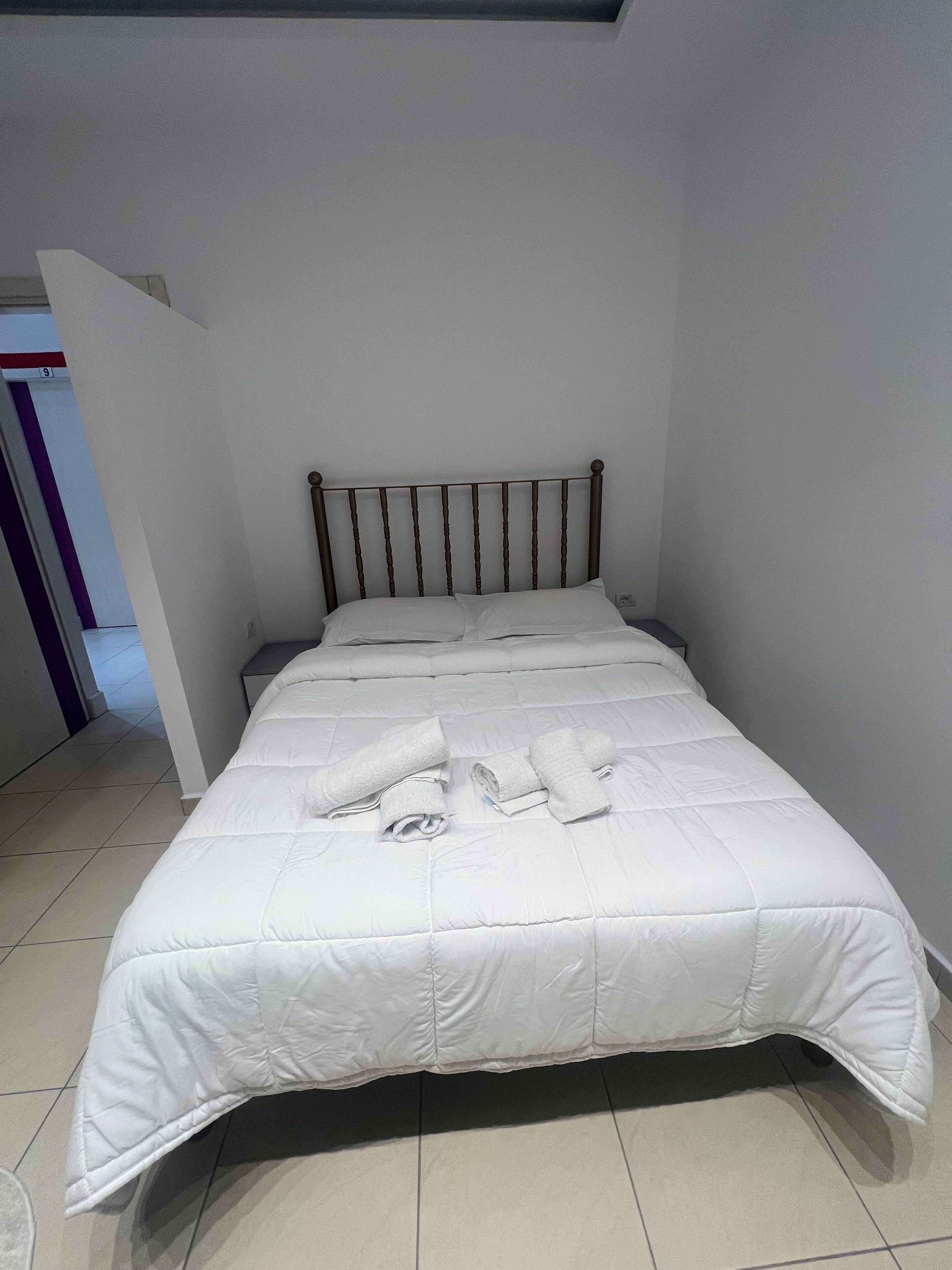 A neatly made bed with a white duvet and rolled towels, a wooden headboard, and a tiled floor.