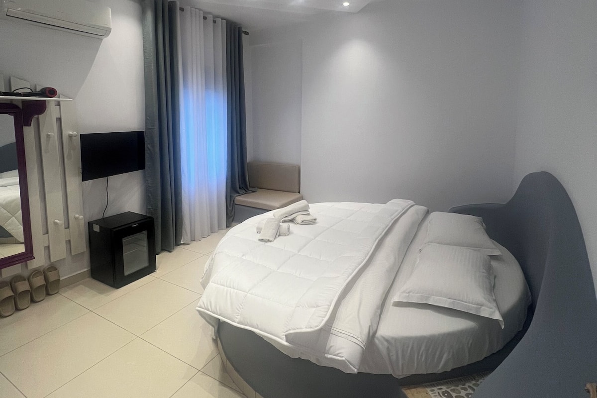 A modern bedroom with a curved bed, a flat-screen TV, a small bedside table, and a built-in air conditioning unit.