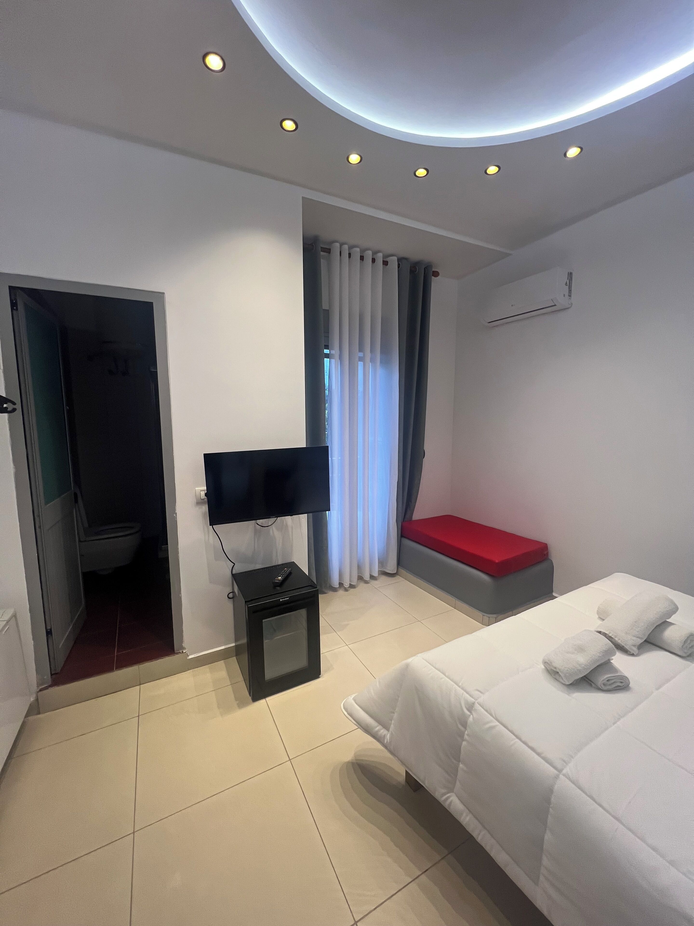 A hotel room with a bed, a TV, a small table, a red bench, and a bathroom visible through an open door.