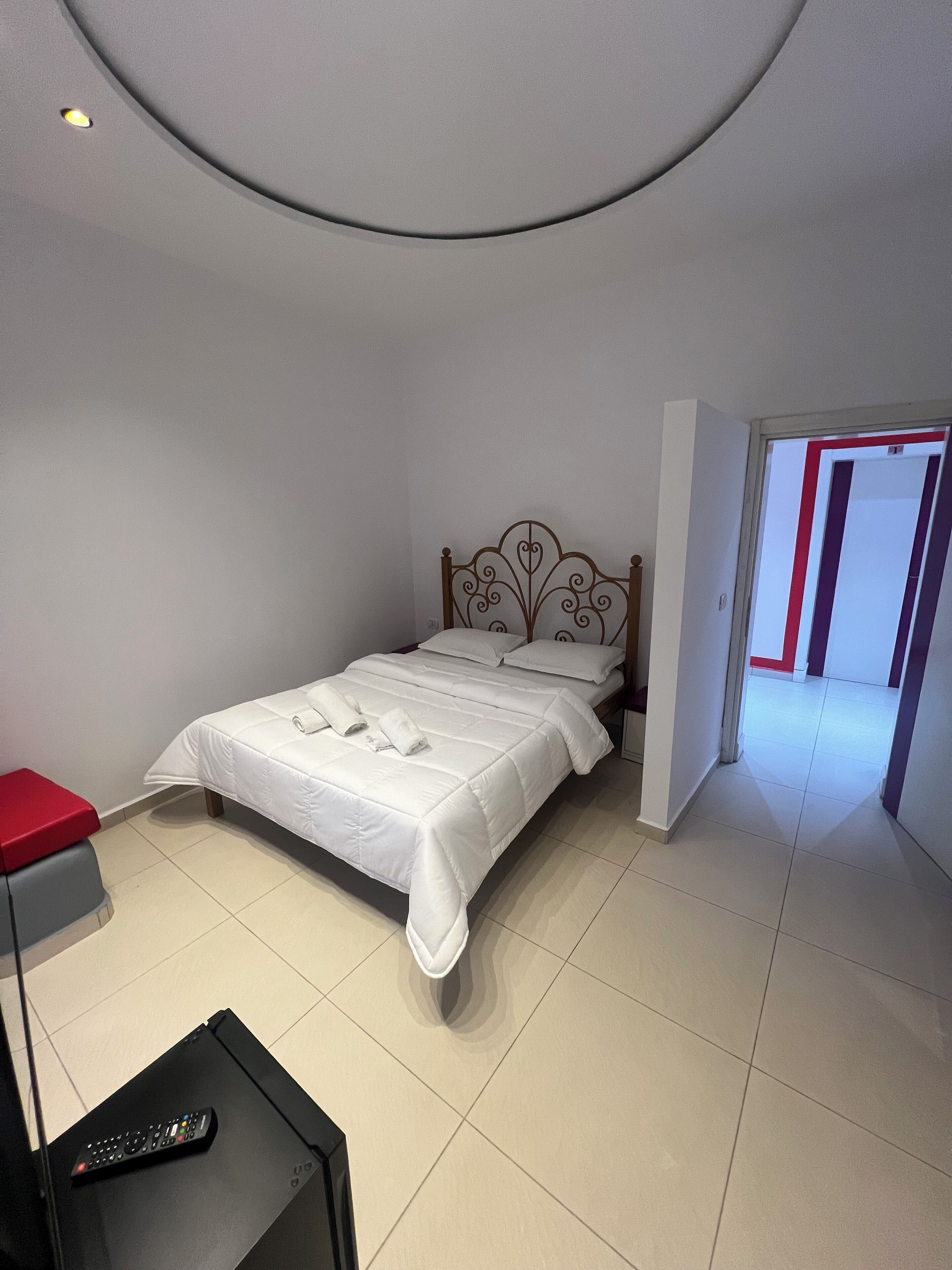 A neatly made bed with white linens and pillows, a red stool, a black table with a remote control, and a door leading to another room.