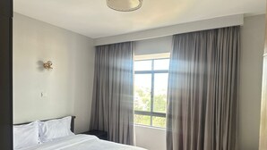 A bedroom with a bed, a window with curtains, a wall-mounted light fixture, and a small bedside table. - Spacious 5-bedroom apartment in terrific Nairobi -White Oak Residency with AC, (Nairobi)