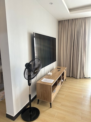 Un salon moderne doté d’un téléviseur à écran plat fixé au mur, d’un ventilateur sur pied, d’un meuble TV en bois et d’une grande fenêtre avec des rideaux.