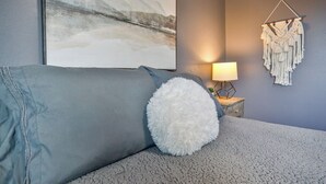 Une chambre avec un canapé gris, un coussin blanc moelleux, une décoration murale en macramé et un tableau représentant un paysage enneigé.