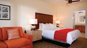 Habitación de hotel con cama, dos mesitas de noche con lámparas, un ventilador de techo, una obra de arte enmarcada en la pared y una colcha roja sobre la cama.