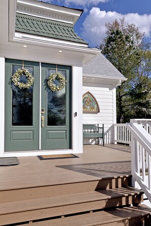 A white building with green doors, a wooden deck, and a bench.