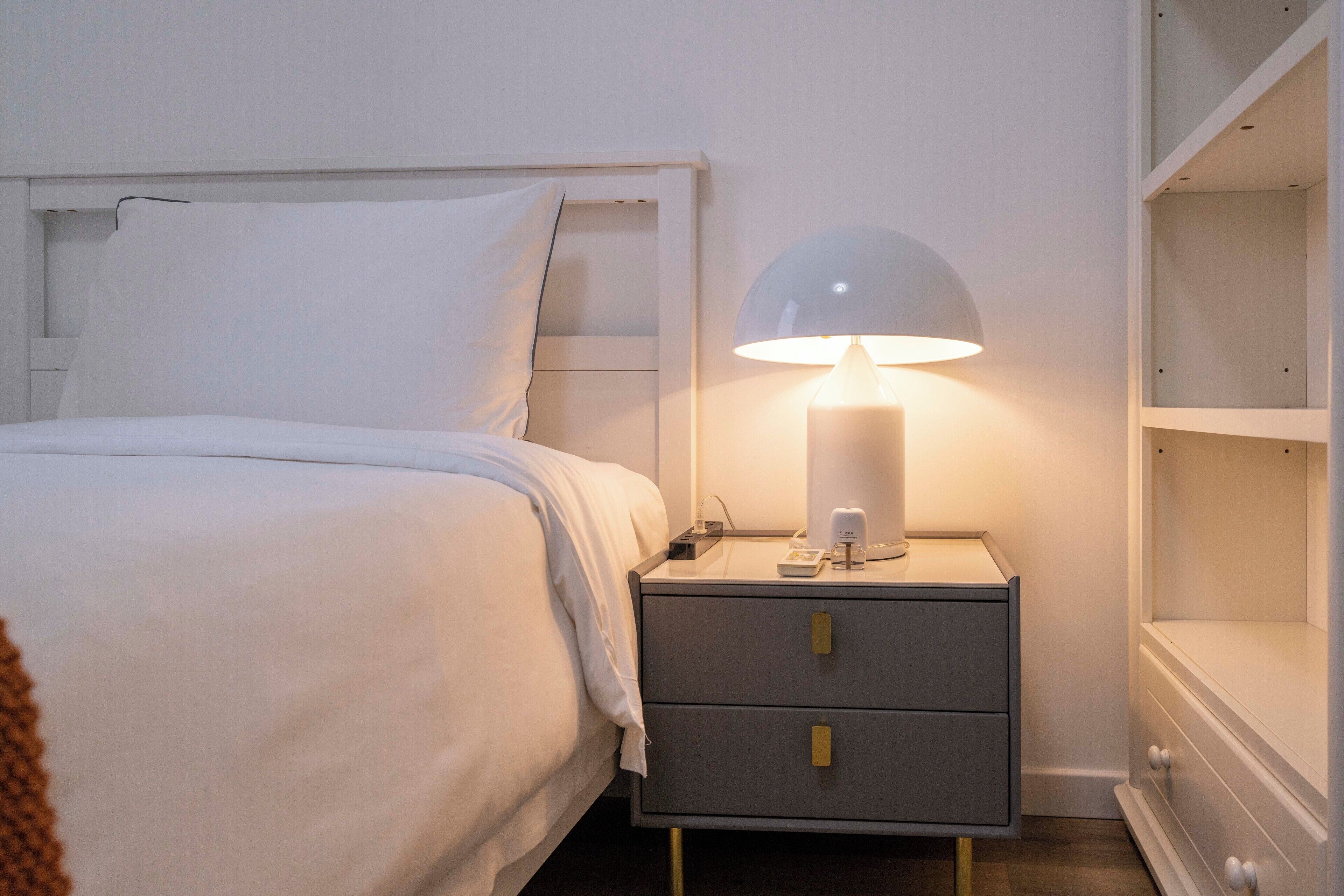 Een netjes opgemaakt bed met wit beddengoed, een bedlampje en een klein nachtkastje met een telefoon en een beker.
