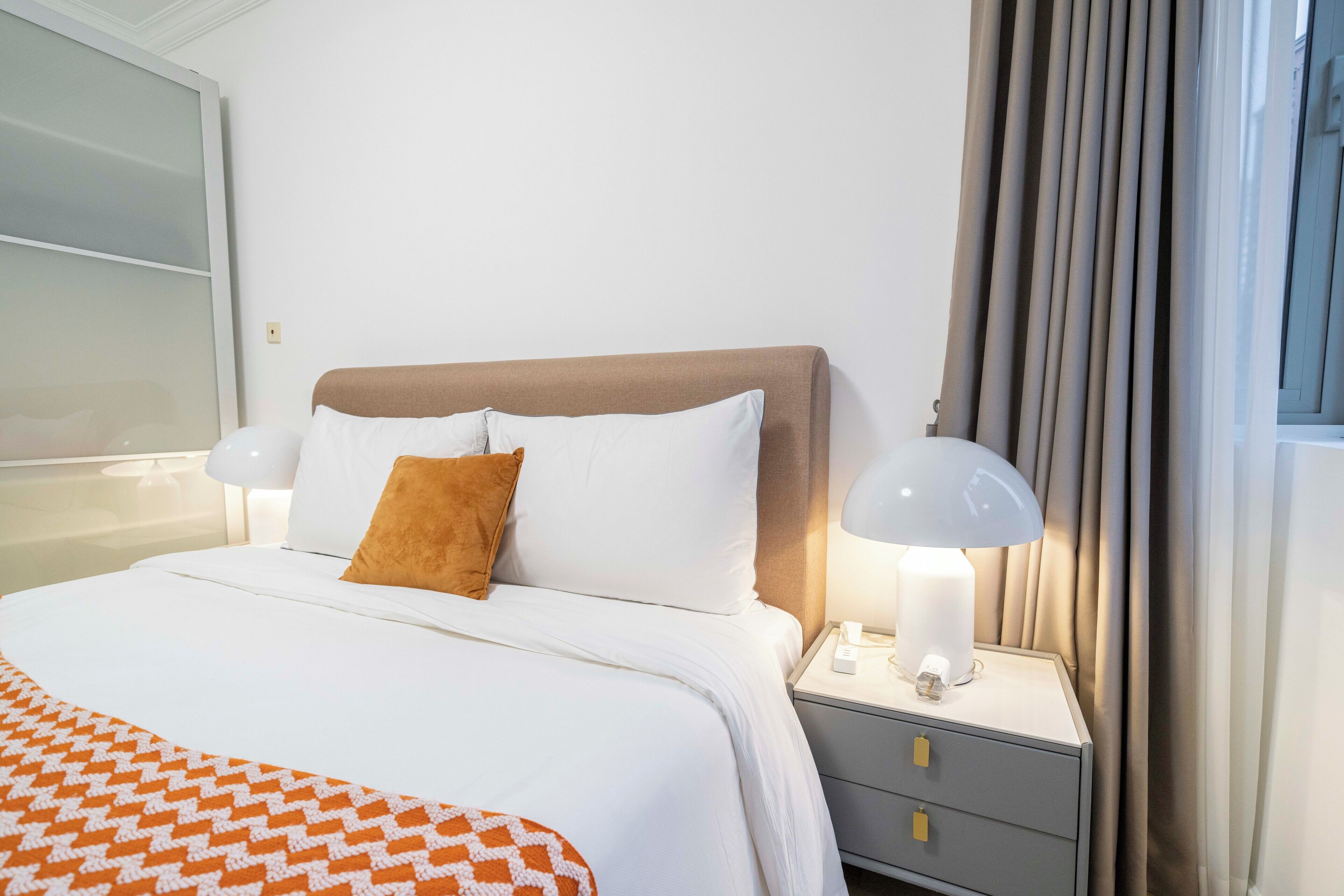Een net opgemaakt bed met wit en oranje beddengoed, een bruin hoofdbord en twee bedlampjes op een grijze nachtkastje met twee lades.