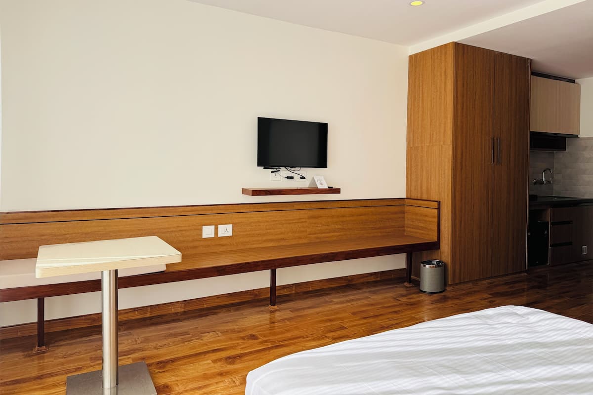 A modern hotel room with a wooden bench, a flat-screen TV mounted on the wall, a small table, and a kitchenette area.
