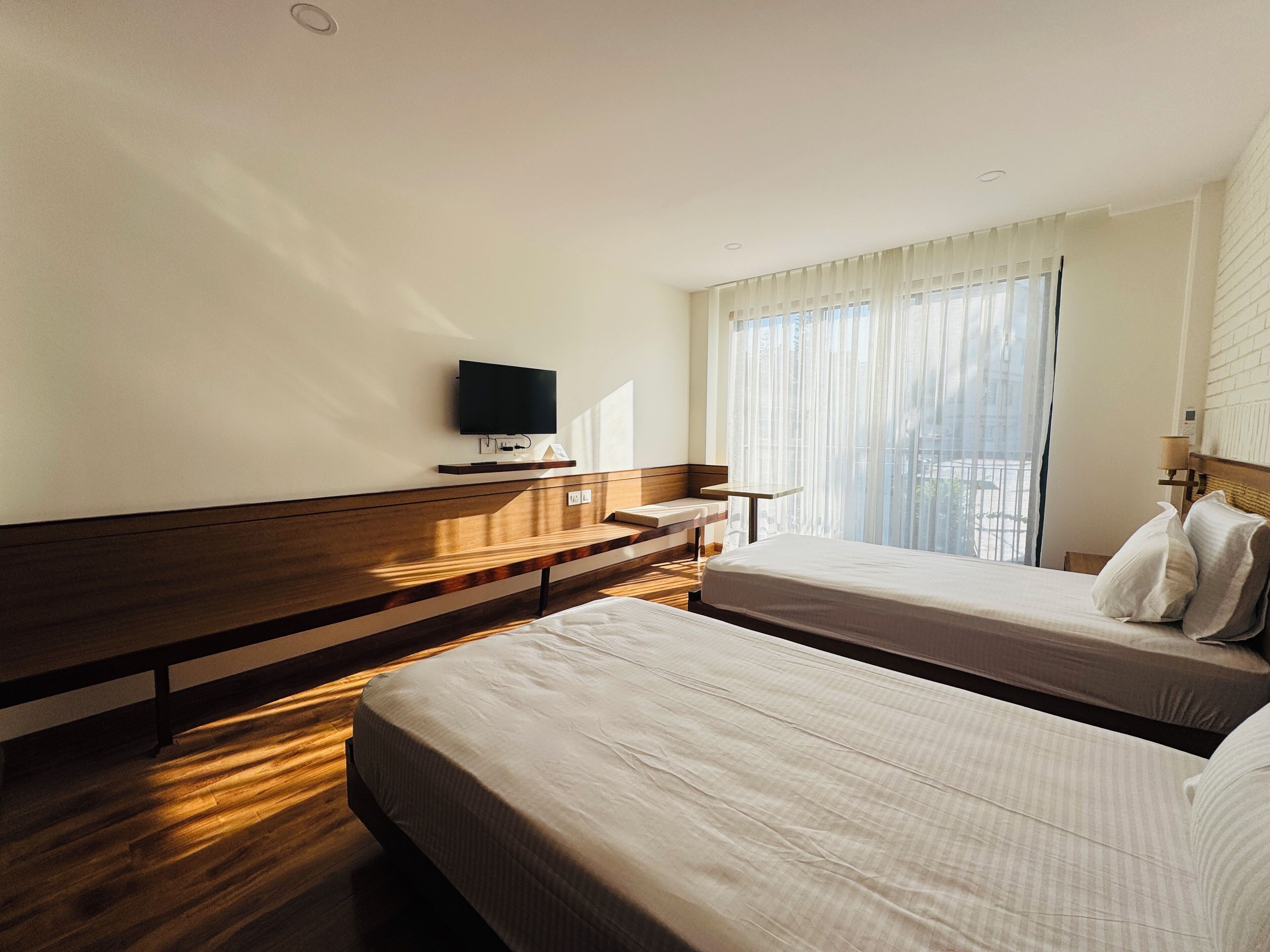 Habitación de hotel con dos camas, un banco de madera, un televisor montado en la pared y un ventanal con cortinas traslúcidas.