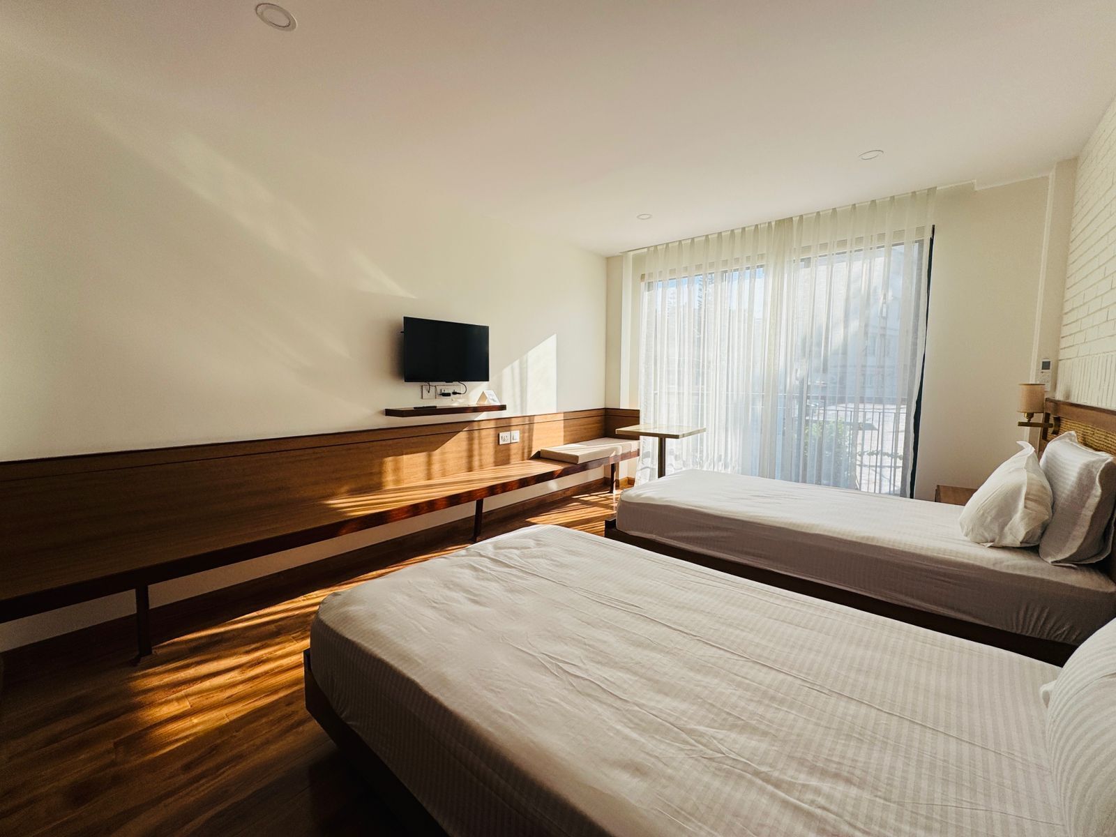 A hotel room with two beds, a TV on the wall, a wooden bench, and a window with curtains.