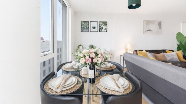 Un espace de salle à manger moderne, avec une table en verre dressée pour quatre personnes, entourée de chaises en cuir noir. On aperçoit, à l’arrière-plan, un canapé gris agrémenté de coussins jaunes.