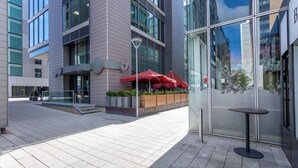 Un bâtiment moderne doté de grandes fenêtres, d’une entrée avec une porte en verre et d’un espace extérieur aménagé avec des parasols rouges.