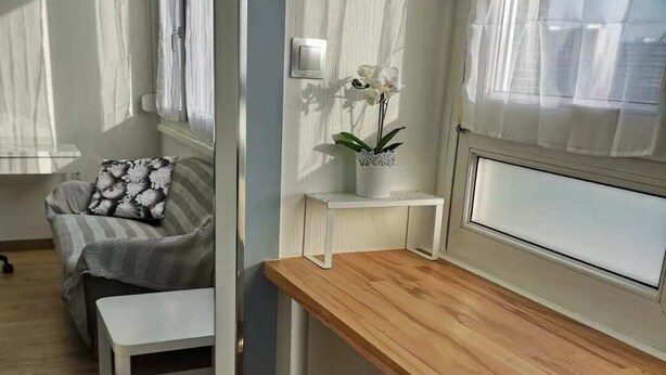 Ein modernes Wohnzimmer mit einem Holztisch, zwei Hocker, einer Couch mit einem gemusterten Kissen und einer Topf-Orchidee.