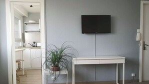 Ein modernes Hotelzimmer mit weißem Schreibtisch, einem Blumentopf, einem flachbildfernseher an der Wand und einer sichtbaren Küchenzeile im Hintergrund.
