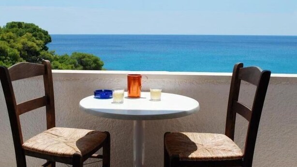 A round table with two chairs, a blue bowl, an orange vase, and two glasses on a balcony overlooking a clear blue sea and green trees. - Three Brothers Hotel (Aegina)