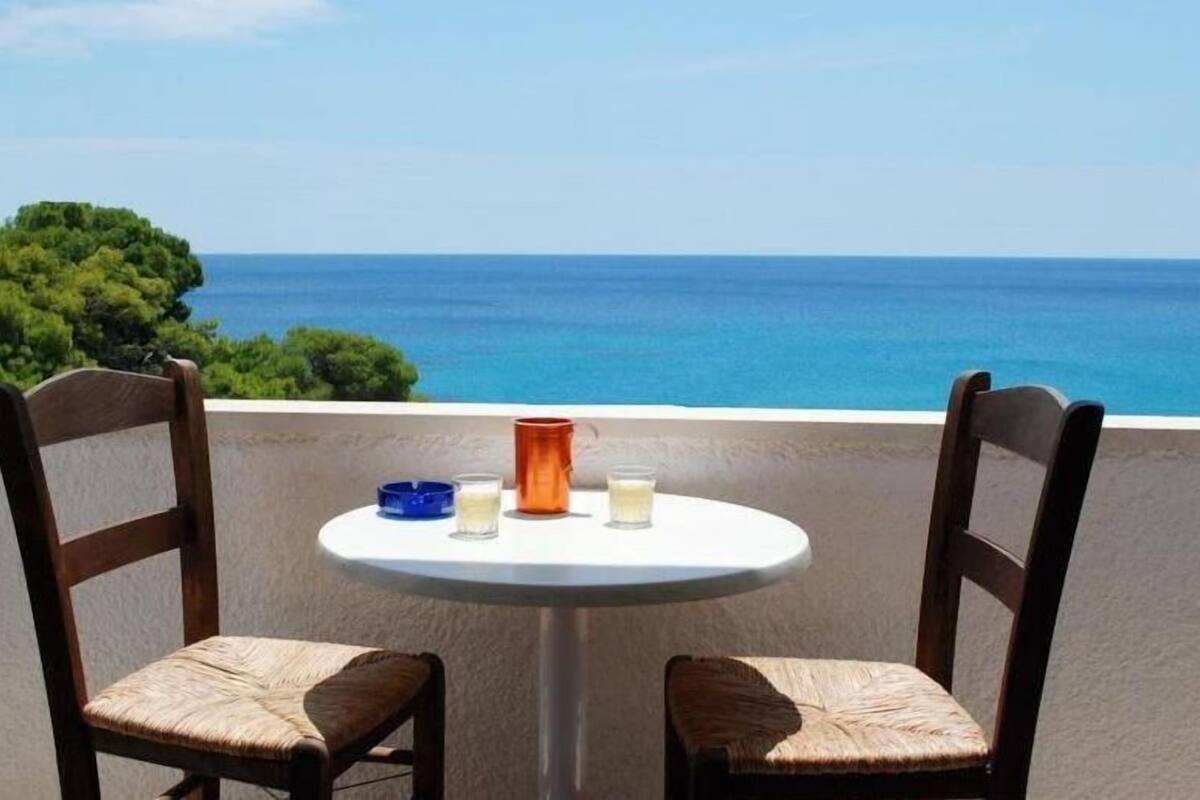 Une table ronde avec deux chaises, un bol bleu, un vase orange et deux verres, le tout placé sur un balcon donnant sur une mer bleue et limpide et sur des arbres verdoyants.