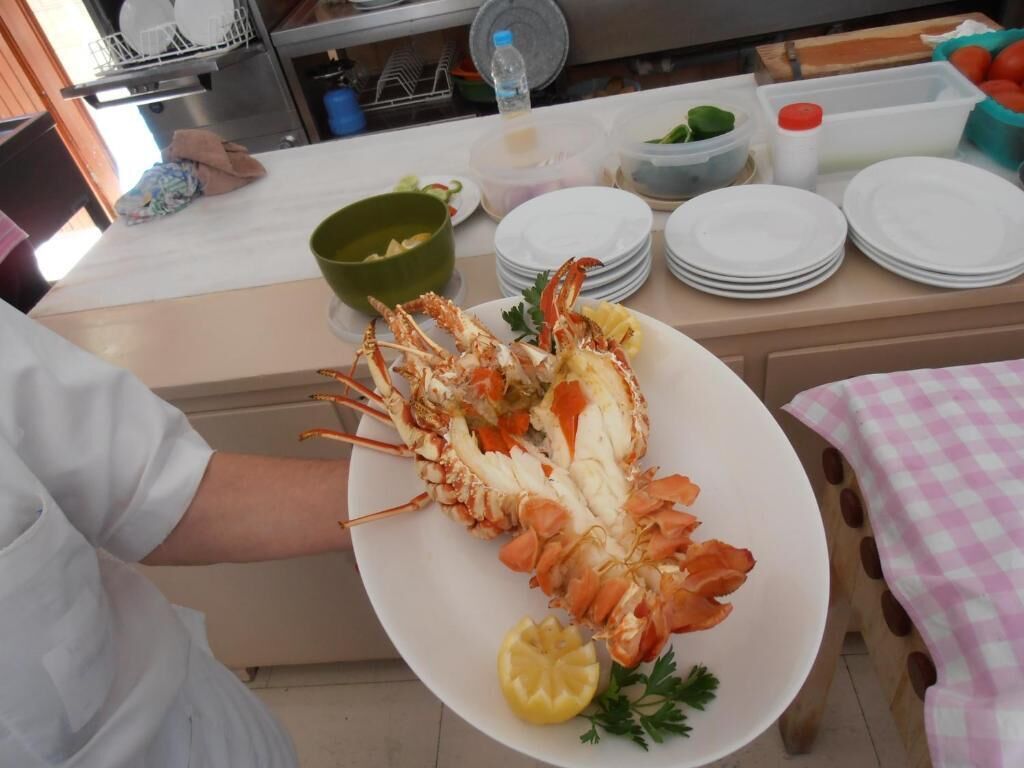 A person holding a large lobster on a white plate, with a checkered tablecloth and kitchen utensils in the background.