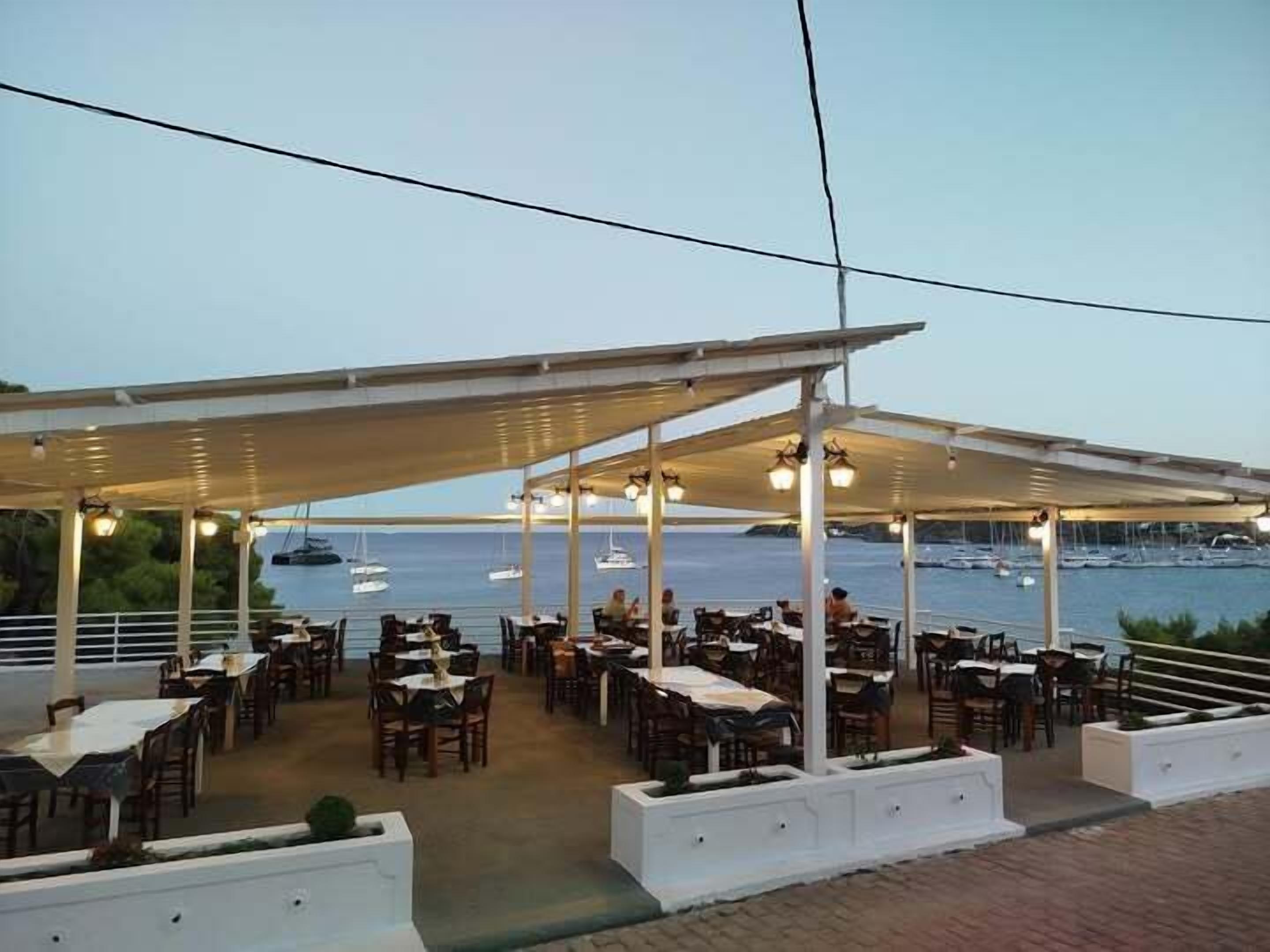 An outdoor dining area with tables and chairs under a covered patio, overlooking a marina with boats.