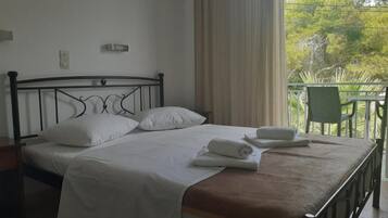 A neatly made bed with white linens and a brown blanket, a green chair, and a view of greenery outside.