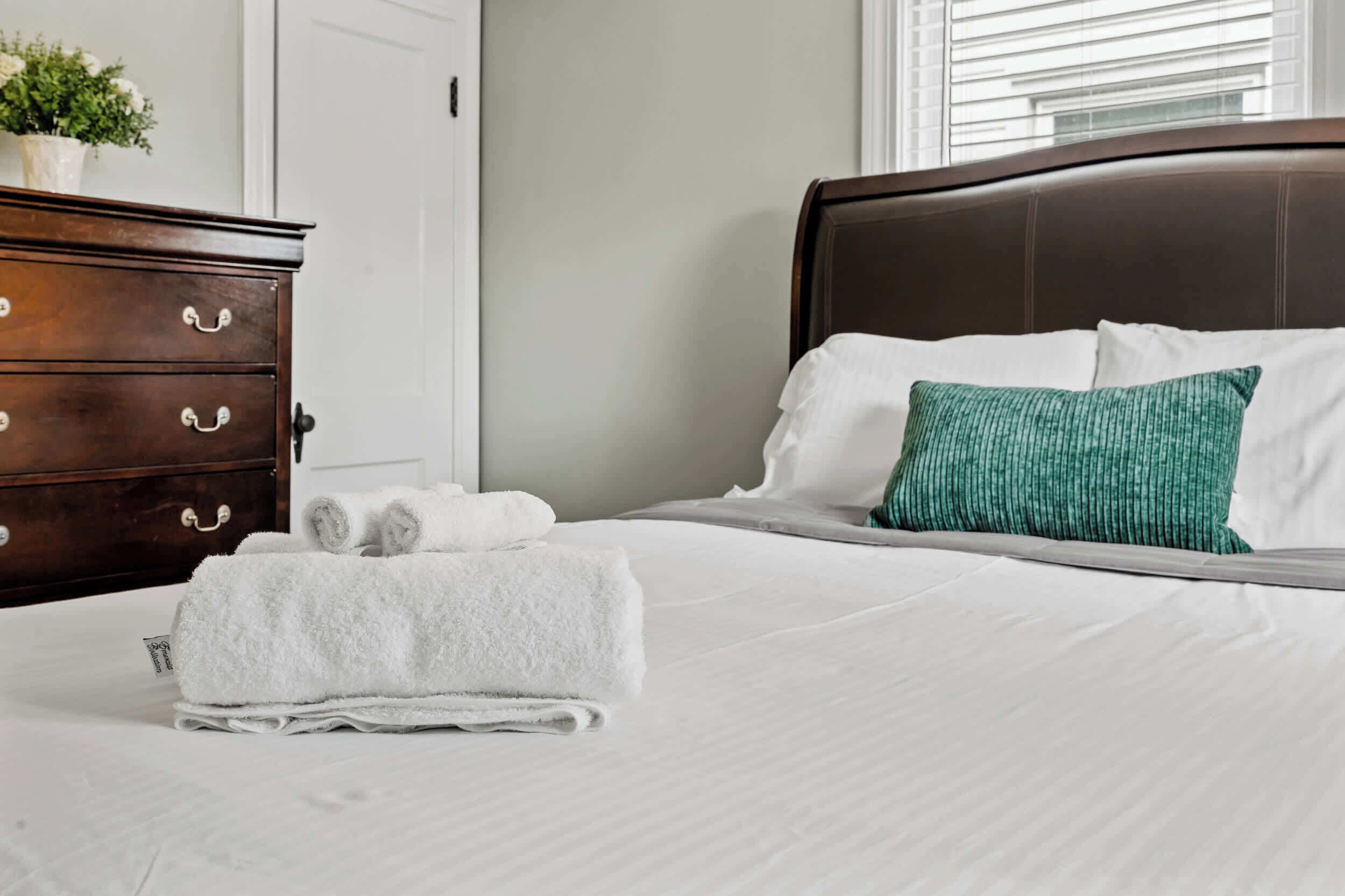 A neatly made bed with white linens and a green pillow, a wooden dresser with drawers, and a potted plant on the dresser.
