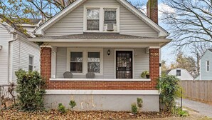 A single-story house with a brick foundation and white siding, featuring a front porch with two chairs and a small potted plant. - Your perfect getaway| 1 mile from Churchill Downs! (Louisville)