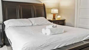 A neatly made bed with white linens and rolled towels, a dark wooden headboard, a bedside table with a lamp, and a white door. - Your perfect getaway| 1 mile from Churchill Downs! (Louisville)