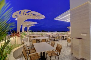 Un área de comedor al aire libre con varias mesas y sillas, rodeada de plantas y estructuras blancas decorativas.