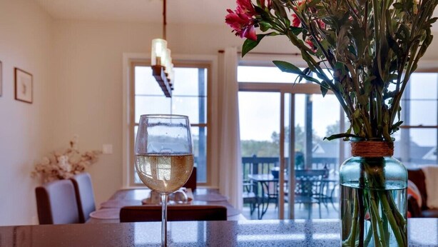 Un verre de vin blanc sur une table, un vase avec des tiges vertes et des fleurs roses, et un coin repas avec des chaises et un banc.